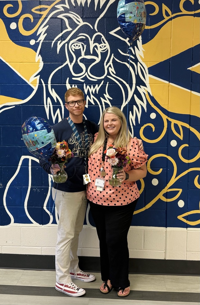 Mr. Rubenstein and Ms. Grant being acknowledged for Teacher of the Year and Support Professional of the Year. A man and a woman stand smiling side-by-side indoors in front of a vibrant blue and yellow mural featuring a stylized white outline of a lion's face. The man, wearing glasses, a dark long-sleeve shirt, and khaki pants, holds a small flower arrangement and a blue balloon. The woman, wearing a pink and black polka-dot blouse and black pants, also holds a small floral arrangement.