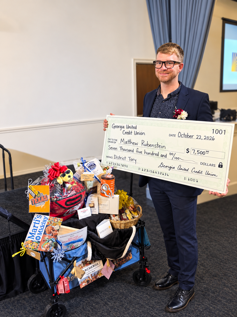 A man in a navy suit, glasses, and a boutonniere smiles while holding a $7,500 check from Georgia United Credit Union. Beside him is a blue wagon filled with gift baskets, snacks, chocolates, and themed gifts at an indoor award ceremony.