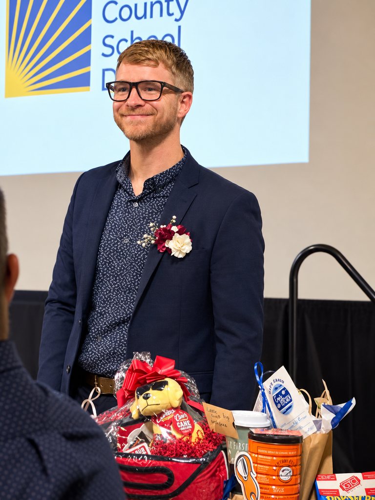 A smiling man wearing glasses, a navy blazer, and a patterned shirt with a burgundy-and-white boutonniere stands at a school recognition ceremony beside a gift basket filled with snacks, a plush mascot, and decorative ribbons. A school presentation slide with a blue-and-gold sunburst logo is projected behind him.