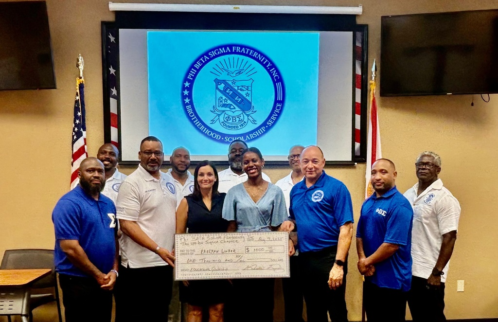 A group photo featuring Raelynn Walker, Becky Murphy, and several members of Phi Beta Sigma Fraternity, Inc., Tau Upsilon Sigma Chapter, all standing in front of a screen displaying the fraternity’s crest. The large $1,000 scholarship check is displayed in front.