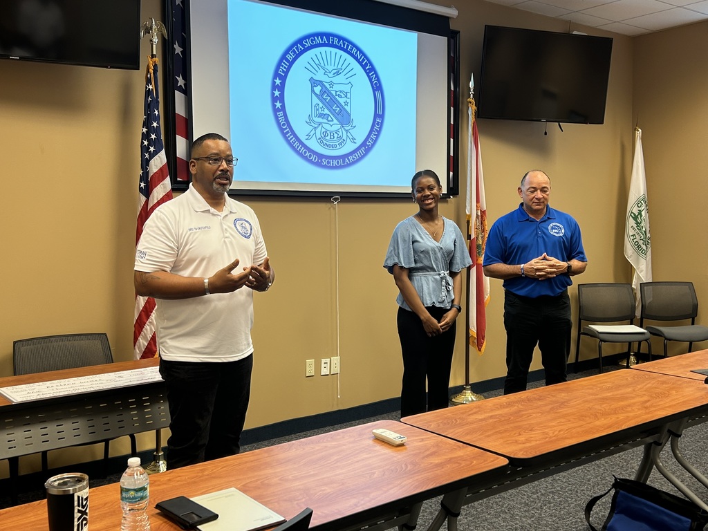 Three people standing at the front of a meeting room — a Phi Beta Sigma Fraternity, Inc. member speaking on the left, Raelynn Walker smiling in the center, and another fraternity member on the right. The fraternity’s crest is displayed on the screen behind them.