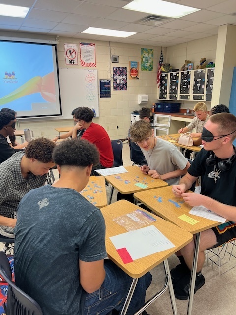 Several high school students work in small groups on jigsaw puzzles. One student in the foreground is blindfolded while participating. Classroom posters and a whiteboard are visible in the background.