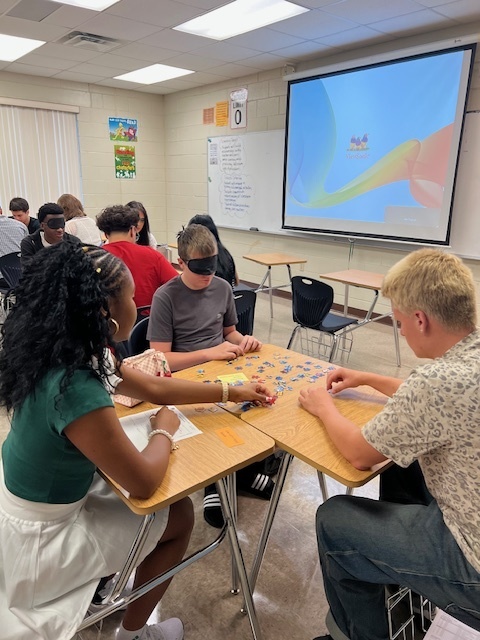 Three high school students sit at desks working on a jigsaw puzzle. One student is blindfolded while the others assist. A projector screen is visible in the background with the “Welcome” slide.