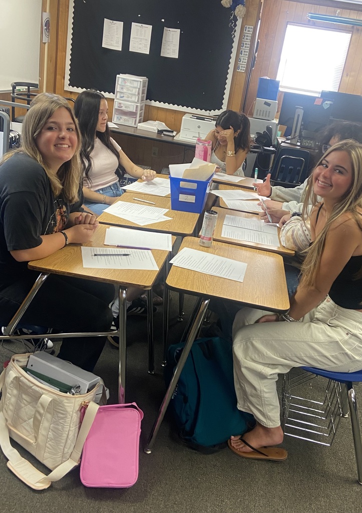 Four high school students sit around a group of desks, smiling and working on a class activity. Papers and pencils are on the desks, and a blue supply bin is in the center.