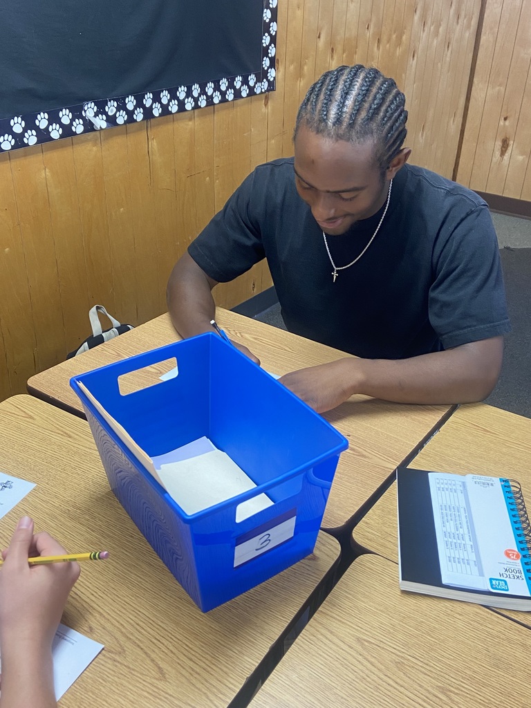 A student is seated at a desk, smiling slightly while looking down at a paper. A blue bin labeled “3” and a notebook are on the desk in front of him.