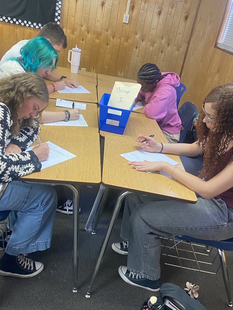Five high school students are focused on writing during a group activity. They are seated around desks with a blue supply bin labeled “Group 1” in the center.