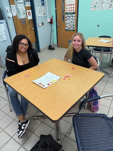 Students sitting in small groups at classroom tables, holding Starburst candies and talking to each other as part of an icebreaker activity.
