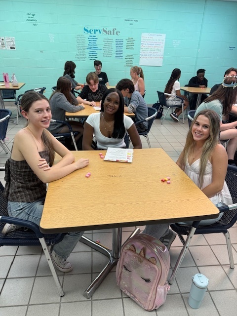 Students sitting in small groups at classroom tables, holding Starburst candies and talking to each other as part of an icebreaker activity.