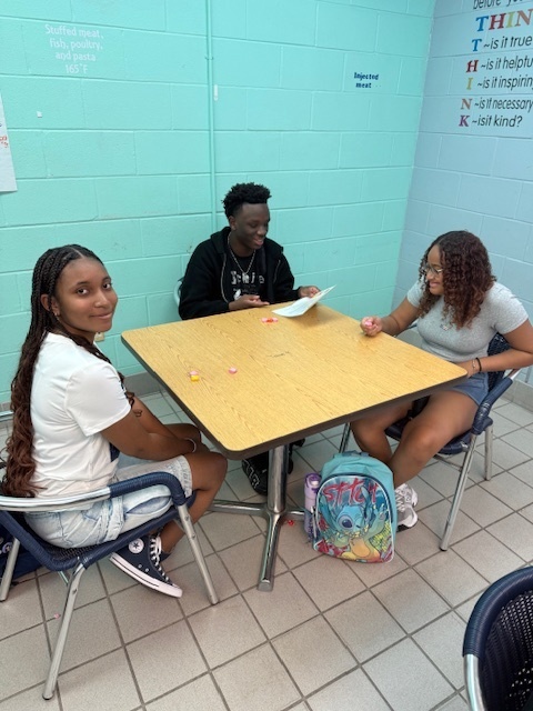 Students sitting in small groups at classroom tables, holding Starburst candies and talking to each other as part of an icebreaker activity.