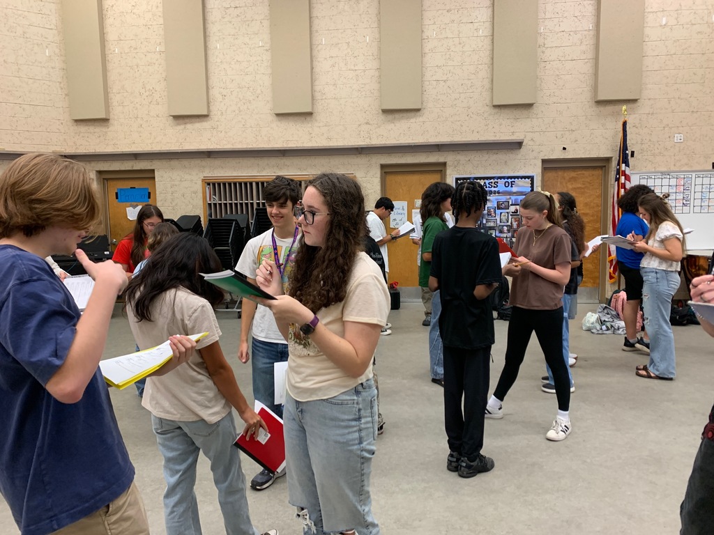Students from Ms. Bice’s and Mr. Colón’s AICE/IB Music, chorus, and band classes sitting and standing in small groups in a classroom, talking and smiling during an icebreaker “get to know each other” activity. Musical instruments and classroom materials are visible in the background.