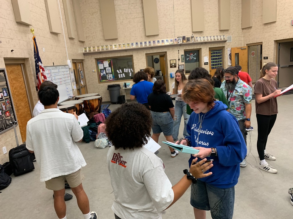 Students from Ms. Bice’s and Mr. Colón’s AICE/IB Music, chorus, and band classes sitting and standing in small groups in a classroom, talking and smiling during an icebreaker “get to know each other” activity. Musical instruments and classroom materials are visible in the background.