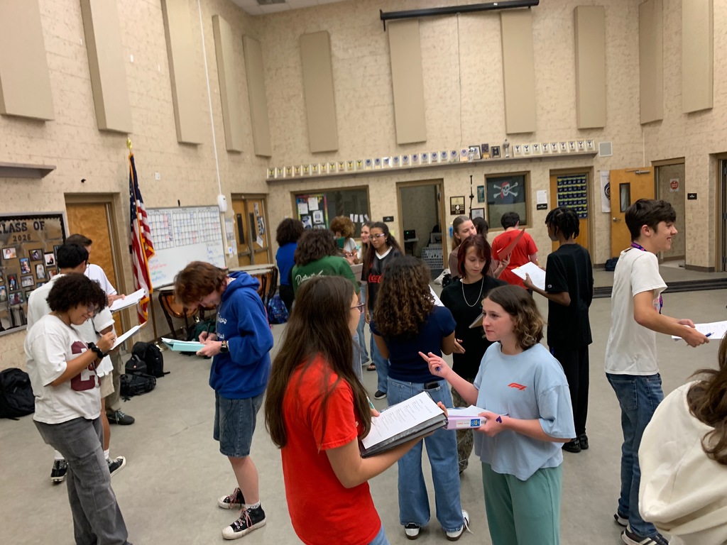 Students from Ms. Bice’s and Mr. Colón’s AICE/IB Music, chorus, and band classes sitting and standing in small groups in a classroom, talking and smiling during an icebreaker “get to know each other” activity. Musical instruments and classroom materials are visible in the background.