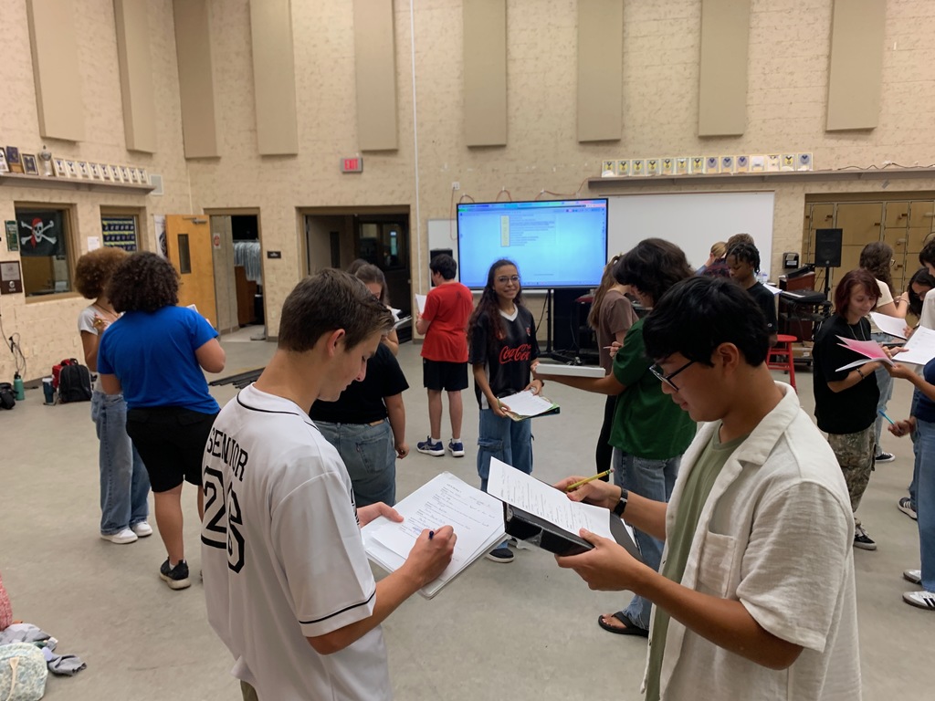 Students from Ms. Bice’s and Mr. Colón’s AICE/IB Music, chorus, and band classes sitting and standing in small groups in a classroom, talking and smiling during an icebreaker “get to know each other” activity. Musical instruments and classroom materials are visible in the background.