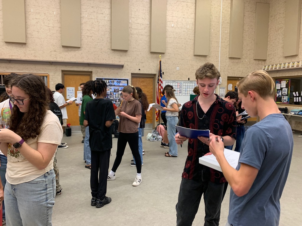 Students from Ms. Bice’s and Mr. Colón’s AICE/IB Music, chorus, and band classes sitting and standing in small groups in a classroom, talking and smiling during an icebreaker “get to know each other” activity. Musical instruments and classroom materials are visible in the background.