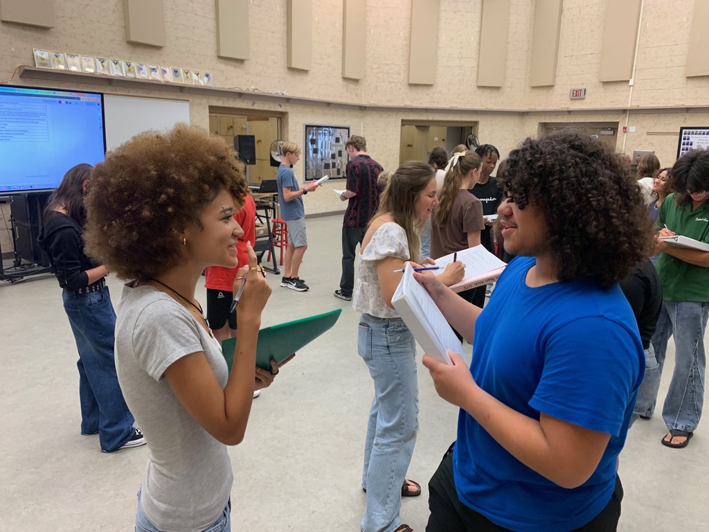 Students from Ms. Bice’s and Mr. Colón’s AICE/IB Music, chorus, and band classes sitting and standing in small groups in a classroom, talking and smiling during an icebreaker “get to know each other” activity. Musical instruments and classroom materials are visible in the background.