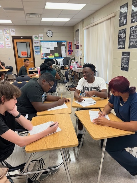 Students in small groups at classroom desks reading, writing, and discussing together during a critical thinking activity in Ms. Holt’s reading class.