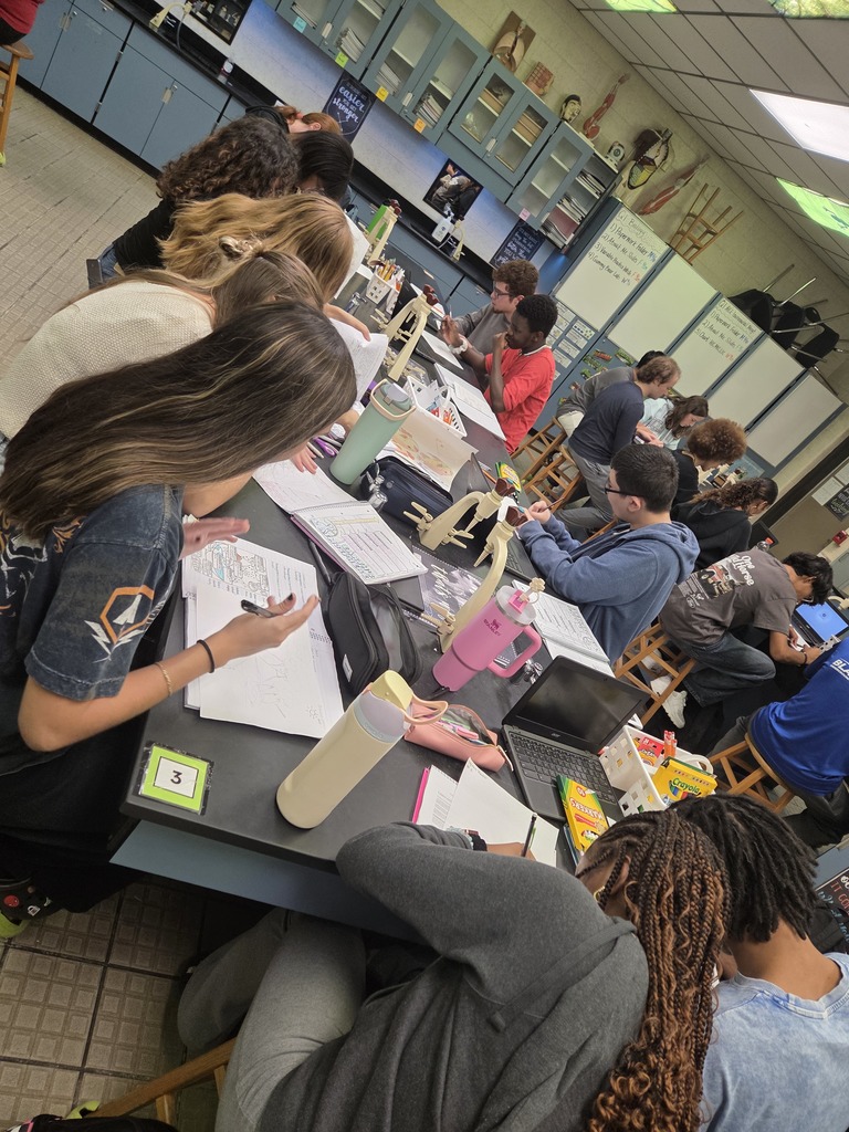 Students working in small groups at classroom desks, using markers and colored pencils to create diagrams on large sheets of paper.