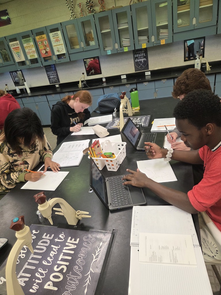 Students working in small groups at classroom desks, using markers and colored pencils to create diagrams on large sheets of paper.