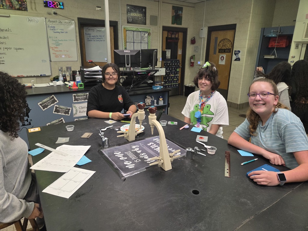 Students in small groups at lab tables measuring gummy bears and recording data in notebooks.