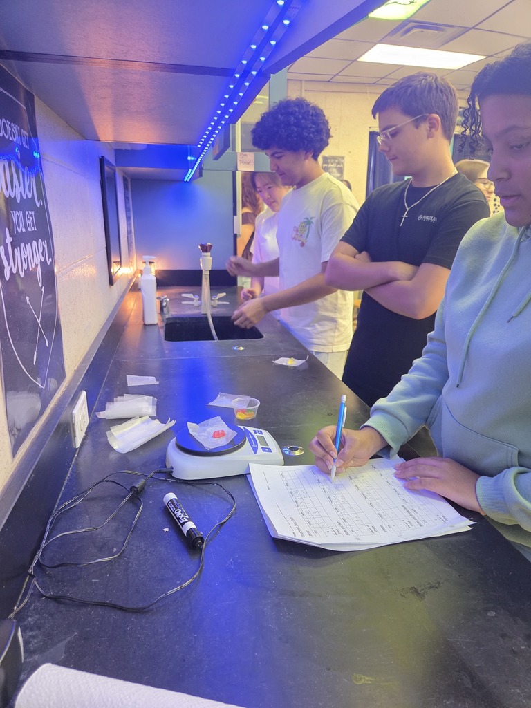 Students in small groups at lab tables measuring gummy bears and recording data in notebooks.
