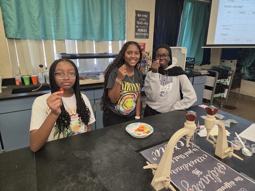 Students in small groups at lab tables measuring gummy bears and recording data in notebooks.