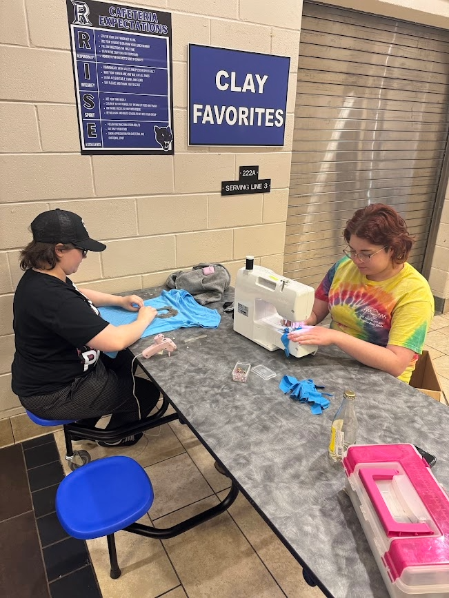 Two theatre students sitting at a cafeteria table using a sewing machine and fabric to prepare costumes.