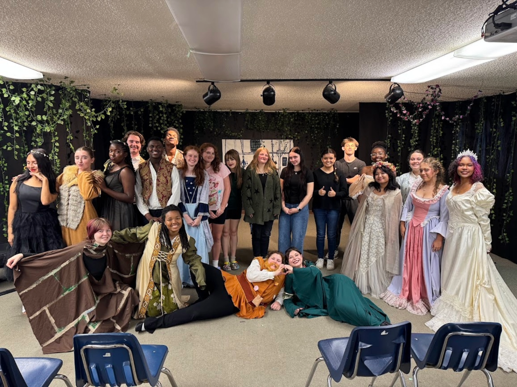 Full theatre class in costume and regular clothes posing together after a performance, with some students wearing elaborate dresses and others in character outfits.