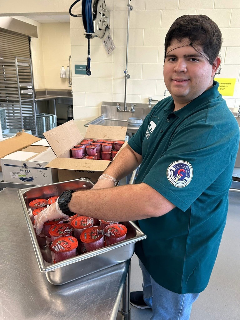 Student in a green Clay County polo shirt places sealed fruit cups into a large metal pan, wearing gloves and smiling at the camera.