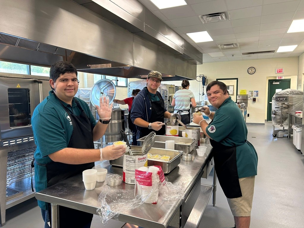 Three students wearing aprons and gloves work together in a school kitchen, smiling while preparing and serving fruit cups at a counter.