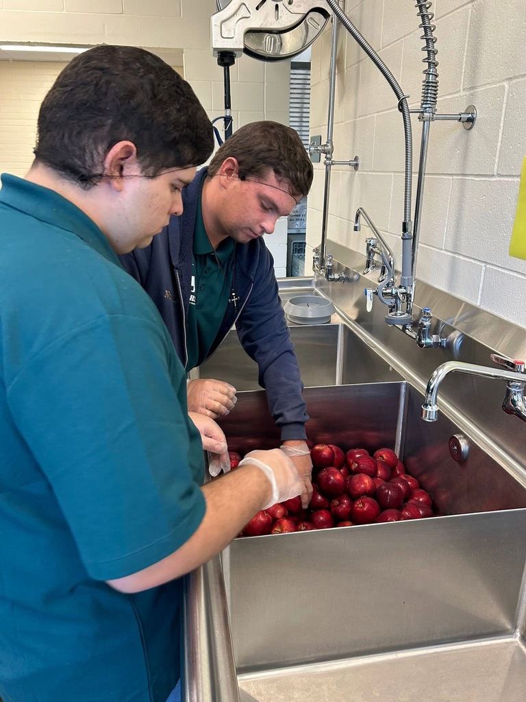 Two students stand at a large sink washing red apples, focused on rinsing and preparing the fruit for use.