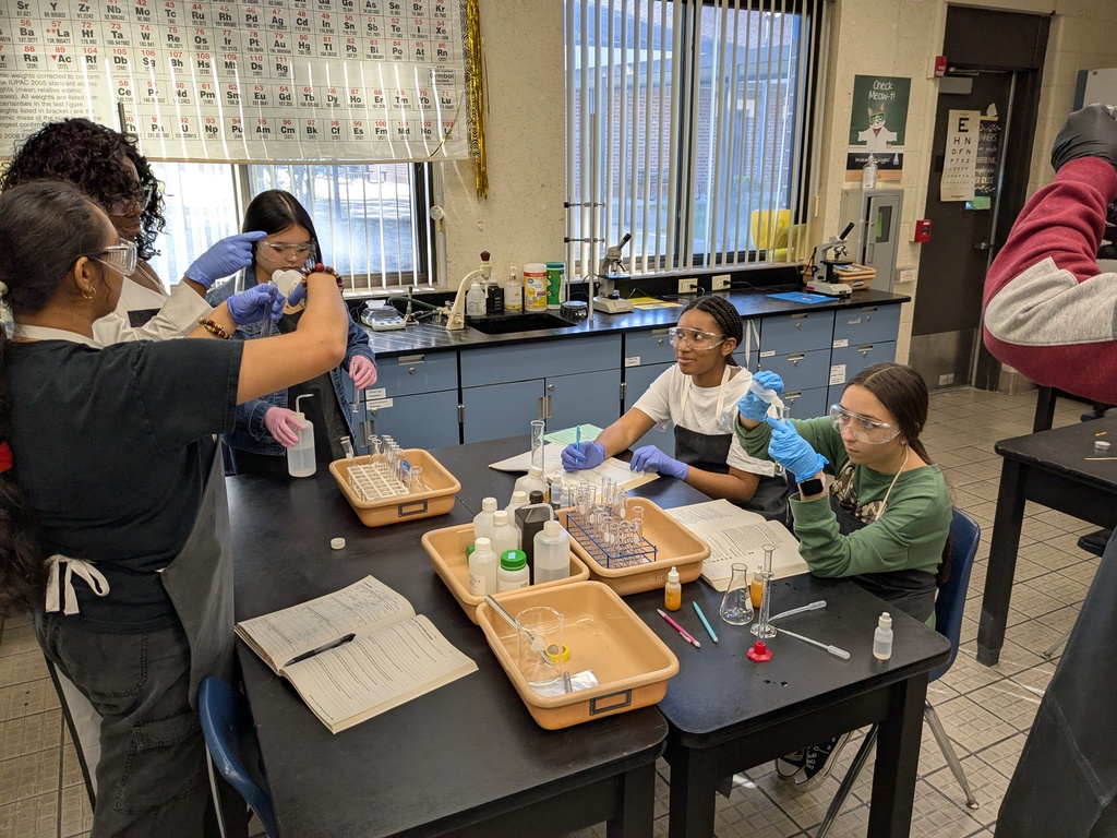 Students in AP/IB Biology wearing safety goggles and gloves work in small groups at lab tables. They handle test tubes, droppers, and trays of lab materials while recording notes in their lab notebooks. A teacher assists one group as they conduct experiments on catalysts and reaction rates.