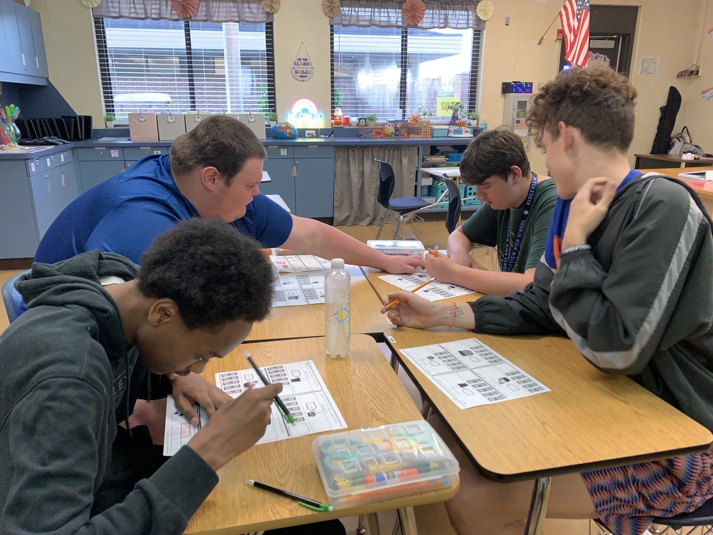 A small group of students sit together at a table, smiling and working with coins and bills during a classroom activity. They are collaborating, discussing, and applying their money skills while their teacher observes nearby.