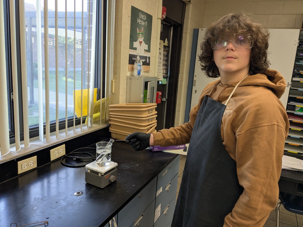 Students in goggles and lab coats measure liquids into test tubes while recording their observations during a chemistry experiment.
