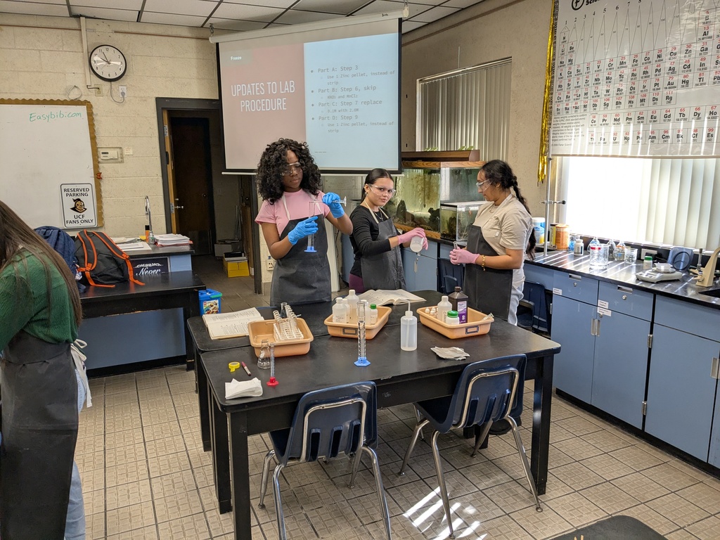 Students in goggles and lab coats measure liquids into test tubes while recording their observations during a chemistry experiment.