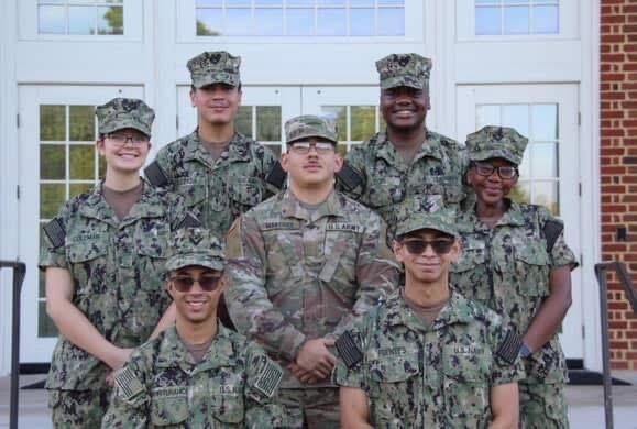 Group photo of seven Ridgeview High School NJROTC alumni standing together in front of a building with white doors and windows. Six are dressed in camouflage Navy uniforms, and one is in an Army uniform. They are smiling and standing in two rows, showing camaraderie and pride.