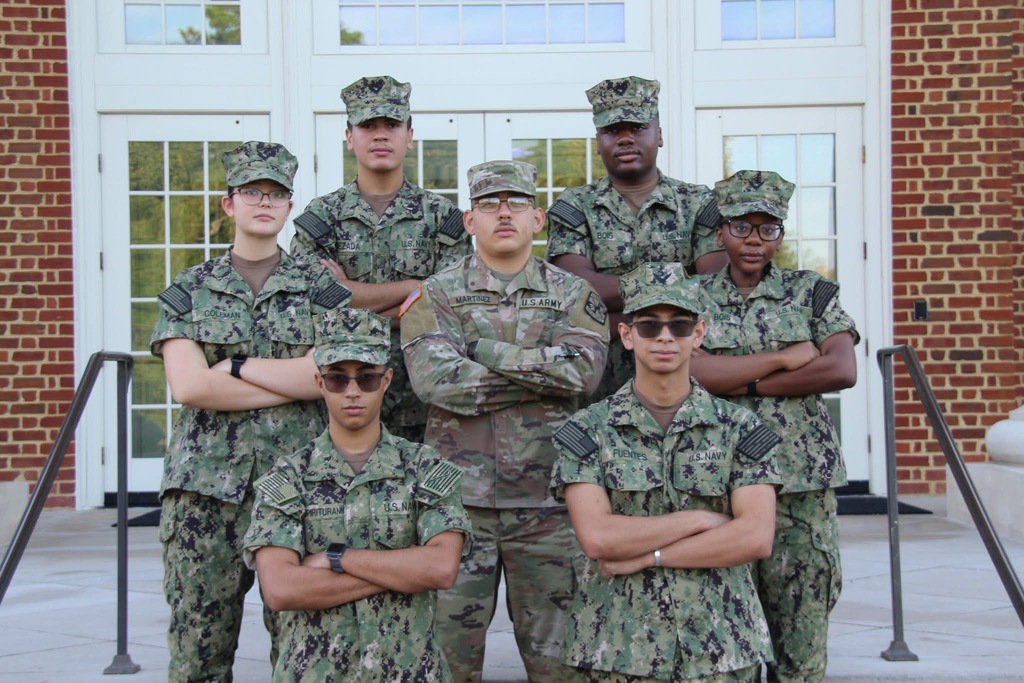 Group photo of seven Ridgeview High School NJROTC alumni standing together in front of a building with white doors and windows. Six are dressed in camouflage Navy uniforms, and one is in an Army uniform. They are smiling and standing in two rows, showing camaraderie and pride.