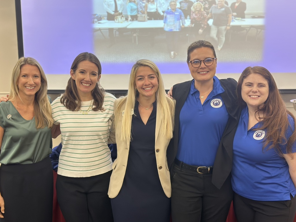A group portrait of five smiling women standing side-by-side in front of a projection screen. From left to right: a woman in a sleeveless olive green blouse, a woman in a green and white striped shirt, a woman in a tan blazer over a dark dress, and two women in matching blue Project REACH polo shirts. The background features a large screen displaying a blurry photo of a larger group of people.