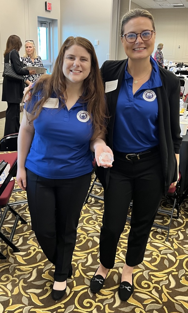 Two women from Project REACH stand together at an awards banquet. Both wear matching royal blue polo shirts with the Project REACH logo and black trousers. The woman on the left has long auburn hair and is smiling broadly while holding a small, clear crystal award in her palm. The woman on the right wears glasses and a black blazer over her polo. They are in a well-lit event hall with circular tables and patterned carpeting in the background.