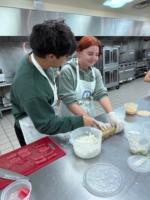 Two people prepare food in a kitchen. One rolls dough, the other mixes ingredients. A table holds various containers.