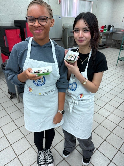 Two women wearing aprons hold food items, smiling. They stand in a kitchen with tiled floors and white walls.