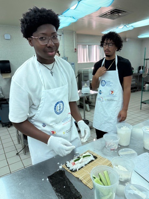 Two people wear aprons and gloves in a kitchen. One prepares food on a table.