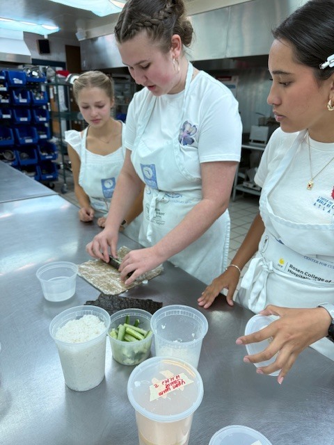 Three women wearing aprons, one with a flower, prepare food in a kitchen. A table holds containers and bowls.