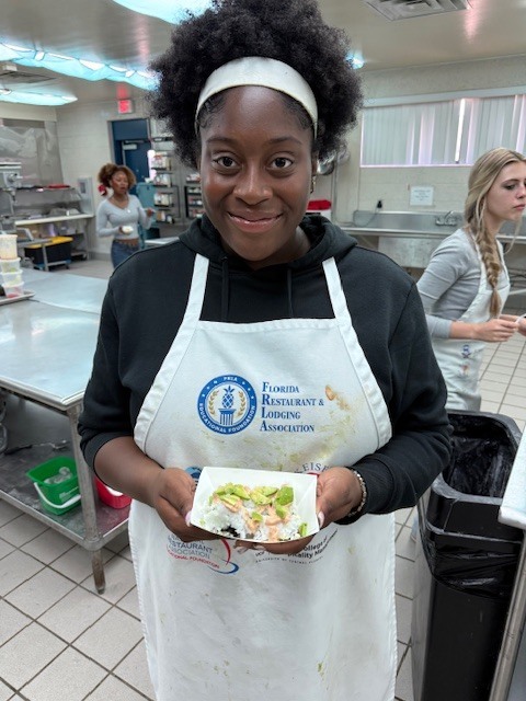 Person in a white apron holding a plate of food in a kitchen. Background shows people and kitchen equipment.