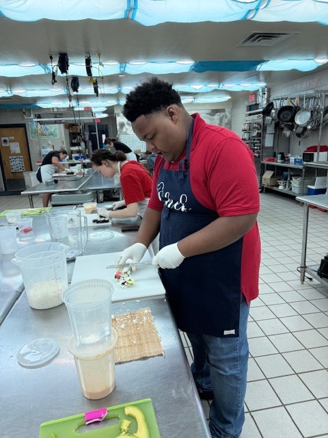 A person wearing a blue apron and red shirt stands in a kitchen, cutting food on a counter.