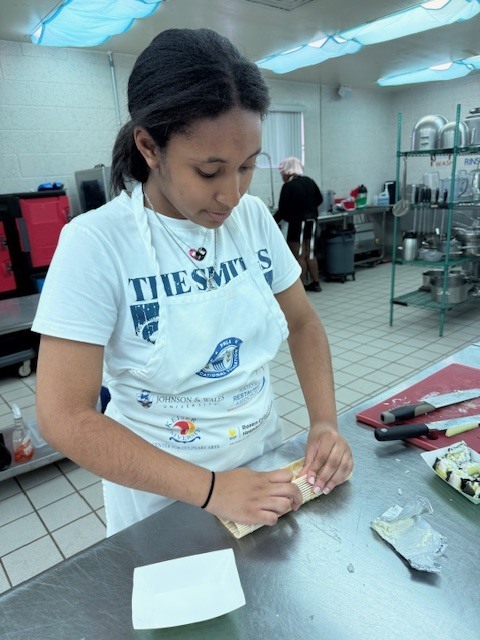 A woman wearing a white apron is standing at a kitchen counter, looking at and preparing food.