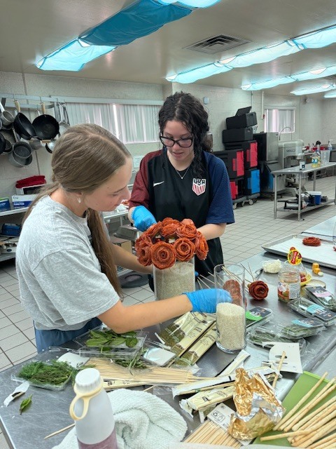 Two individuals in a kitchen, working on a floral arrangement with various materials on a table.