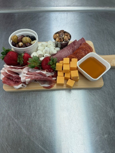 A wooden board displays various foods: strawberries, cheese, cured meats, nuts, olives, and honey, on a metallic surface.