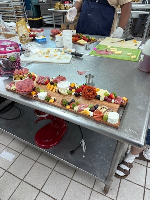 A person in a blue apron prepares a charcuterie board with various cheeses, meats, and fruits.
