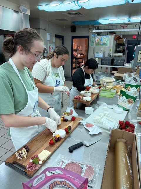 Three people in aprons and gloves prepare food in a kitchen. A table has sliced fruit, cheese, and other items.