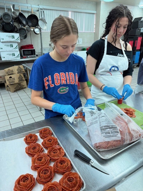 Two people in aprons and gloves prepare food in a kitchen. One person cuts a roll while the other works with meat.